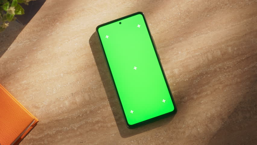 Top View of a Mobile Phone with Vertical Mock Up Green Screen Chromakey Display with Motion Trackers Lying on a Wooden Desk Next to Notebook Organizer with Markers and Take Away Cup of Coffee Royalty-Free Stock Footage #1107300931