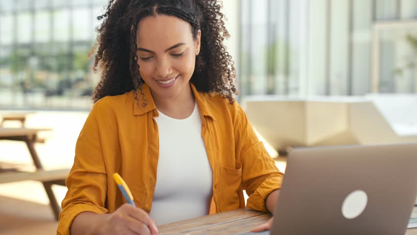 Smart curly brazilian or latino girl, successful female freelancer or student, dressed in an orange shirt, uses laptop to work or study online while sitting outdoors, takes notes in notebook, smile Royalty-Free Stock Footage #1107301283