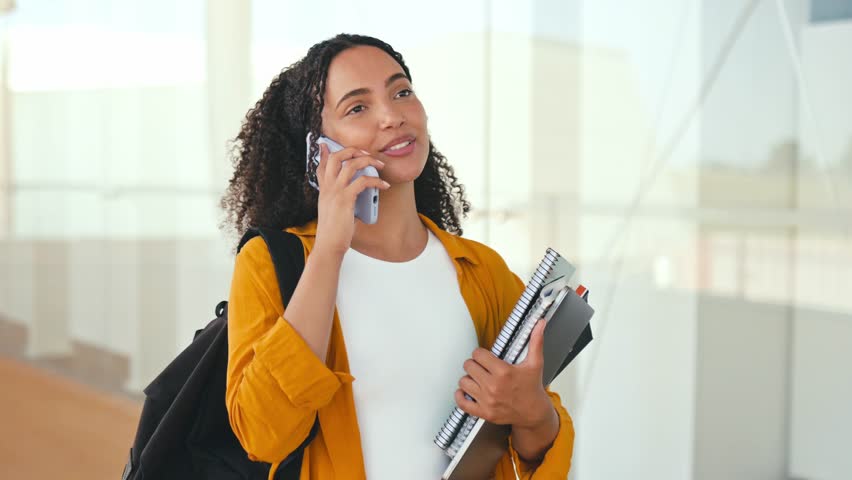 Happy lovely curly haired hispanic or brazilian female student, with a backpack, hold books and notebooks in her hand, stand near the university, talk by smartphone with someone, looking away, smile