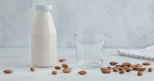 Pouring almond milk into a glass from jug, alternative milk drink made of almond nuts, kitchen table, bottle with no label, vegan drink, white background, slow motion video, horizontal 4k footage