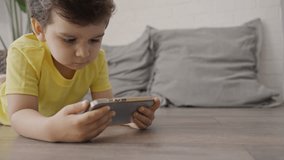 Cute little child boy using smart phone looking at mobile screen, watching cartoons, playing online games, lying on the floor indoors.