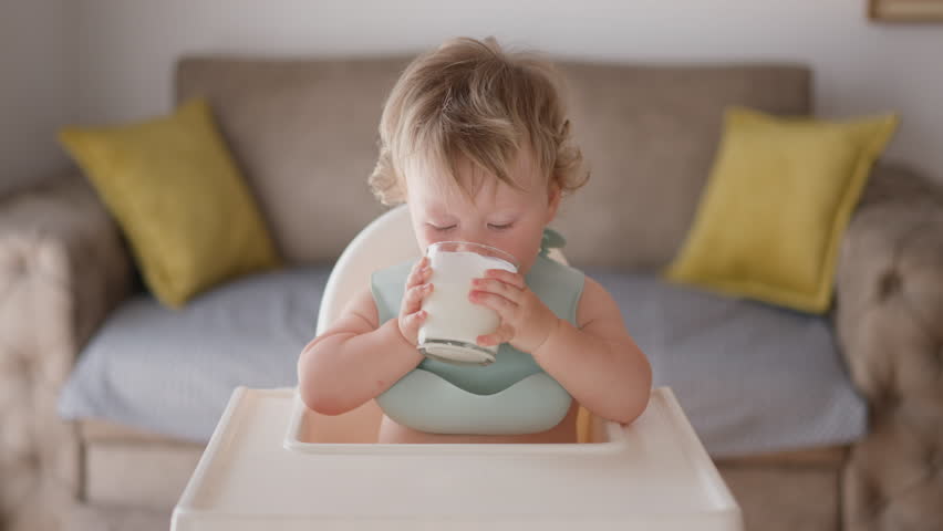 One-Year-Old Sips Nutritious Milk from a Cup: A Healthy Start to Life. little mixed race boy child drinking milk yogurt. Healthy eating toddler having breakfast in living room. Daughter girl holding Royalty-Free Stock Footage #1107320107