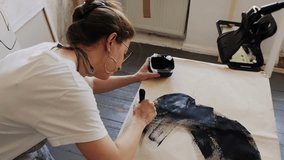 A young artist makes her first strokes on a blank canvas with a large brush and captures it on a camera for a social media. Digital art, artist content. Contemporary art, artist's workshop.