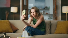 Carefree woman showing tongue to smartphone camera at home. Happy lady making funny face laughing at mobile phone video call. Positive girl holding telephone having fun at weekend apartment alone 