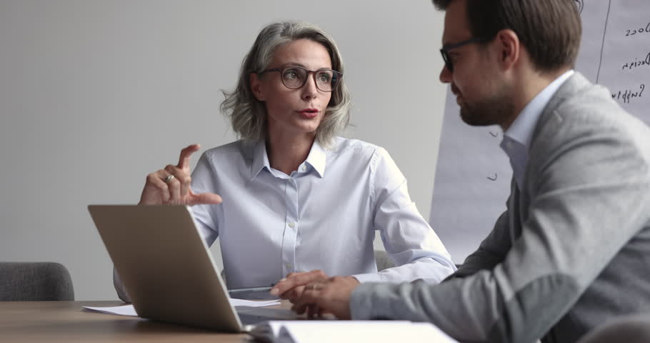Confident beautiful mature business professional woman in glasses talking to younger male coworker at laptop, explaining work strategy, ideas for project, speaking with hand gesture for expression Royalty-Free Stock Footage #1107331677