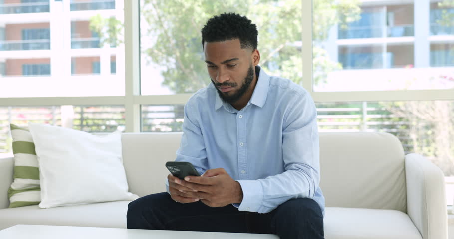 African man sit on sofa with cellphone freaking out, nervous reading bad news in a message feels frustrated get awful sms, personal or business concerns, gadget problems, unexpected badly information Royalty-Free Stock Footage #1107332985