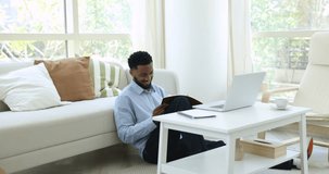 Handsome African man listen webinar, take part in webcast take notes in notebook seated on floor in cozy living room, modern tech usage to gain knowledge, studying, learn program, receive new skills