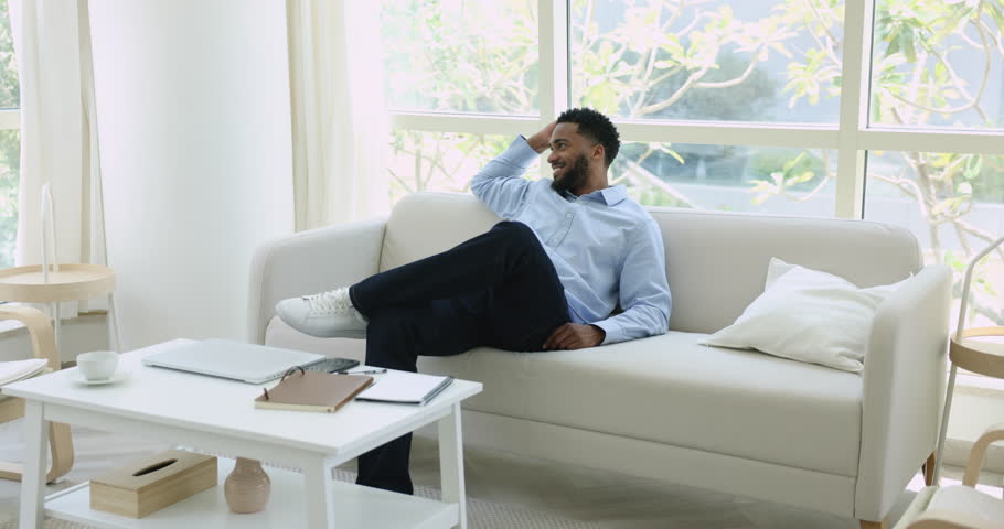 Satisfied African male freelancer finished work relaxing on sofa at homeoffice, smile, daydreaming, looks aside deep in pleasant thoughts feels good with project accomplishment spend free time at home Royalty-Free Stock Footage #1107333001