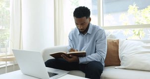 Focused African businessman working from home talk to colleague using video call app and laptop, holds personal organizer write information, takes notes at on-line consultation. Workflow, modern tech