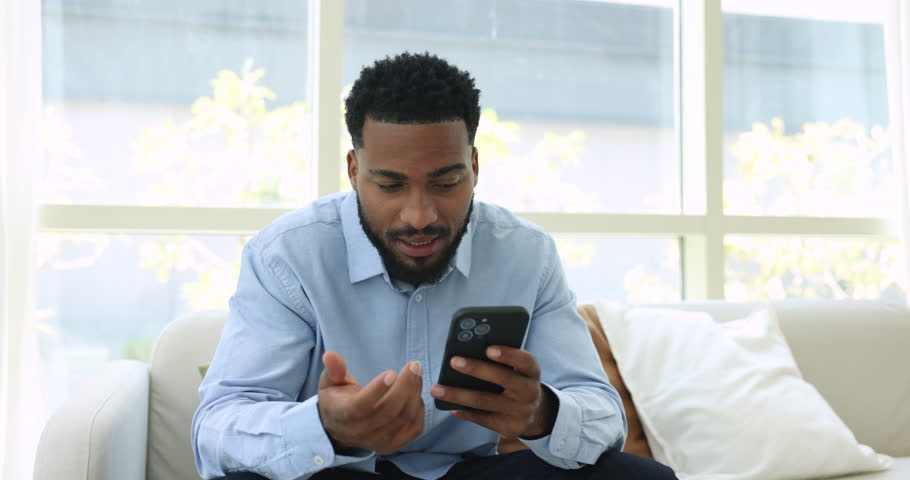 African man holds cellphone talk to friend on speakerphone seated on sofa, lead formal conversation, explain meeting details, make call to customers services, get or give consultation use modern tech Royalty-Free Stock Footage #1107333011