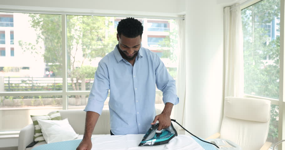 Handsome single African man in casual blue shirt standing alone in modern living room ironing white bath towels on board, smile, doing household daily routine, helps with everyday chores. Housekeeping Royalty-Free Stock Footage #1107336199