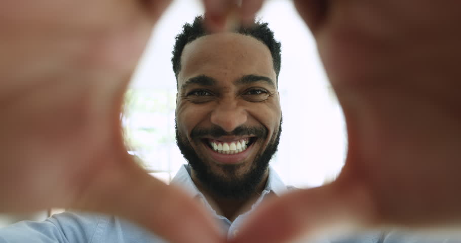 Close up head shot portrait happy handsome African man showing heart symbol smile look at camera through fingers makes sign of love, express kindness, support, solidarity, gratitude, feeling affection