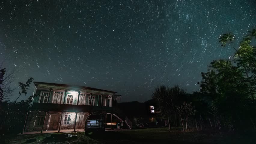 Night sky timelapse with stars trail over village homes showcasing the wonders of the universe. Dark mountains and cosmic delights illuminate the countryside. Astronomical wonders of the galaxy