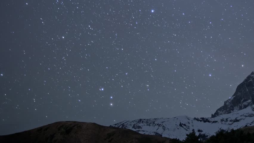 Night with bright stars moving in sky over Mountain Ushba with snowy peaks in time lapse. Landscape of amazing caucasus cliffs and constellations which can be viewed in winter evening during trip