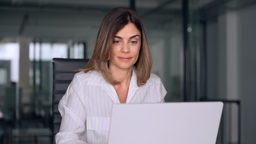 40s mid age European business woman CEO using laptop for work sitting at table in office and looking at camera. Smiling Latin Hispanic mature adult professional businesswoman using pc digital computer Royalty-Free Stock Footage #1107342057