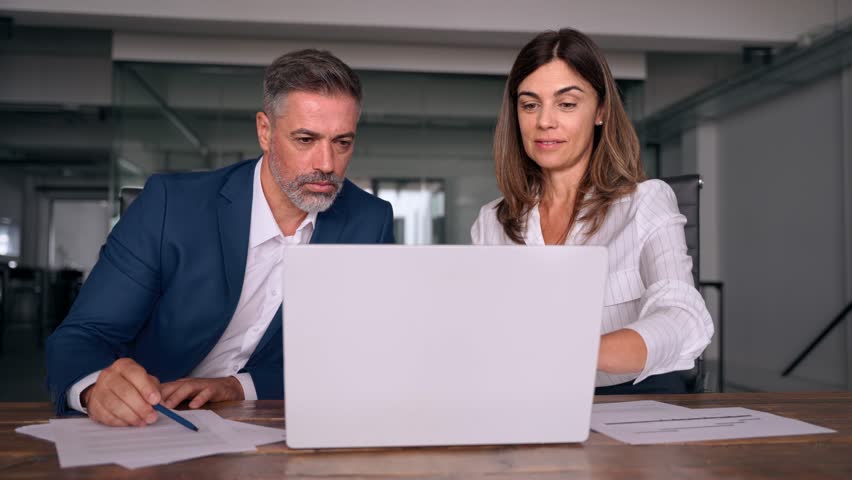 Mature 50s age Latin business man mentoring mid age European business woman discussing project on laptop in office. Two colleagues, group of partners, professional business people working together. Royalty-Free Stock Footage #1107342063
