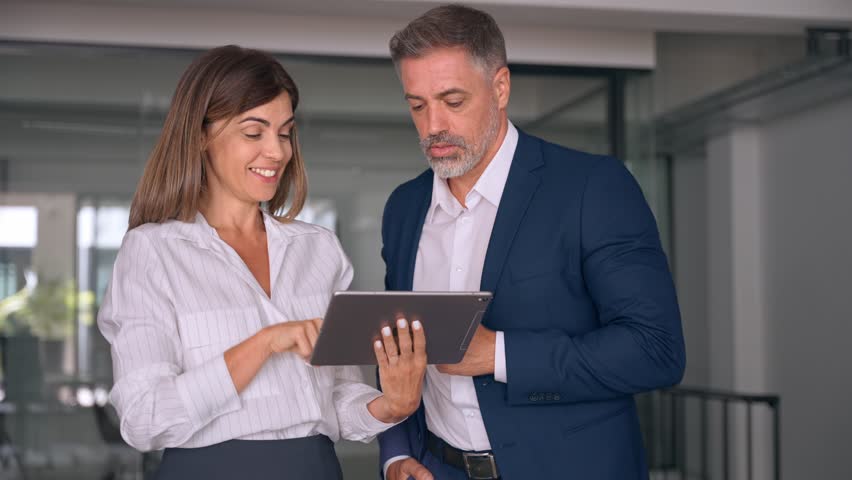 Two diverse partners walking mature Latin business man and European business woman discussing project on tablet in office. Two colleagues of professional business people working together at workspace. Royalty-Free Stock Footage #1107342071