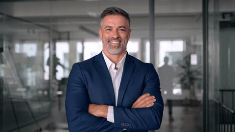 Handsome hispanic senior business man with crossed arms smiling at camera. Indian or latin confident mature good looking middle age leader male businessman on blur office background with copy space. Stockvideó