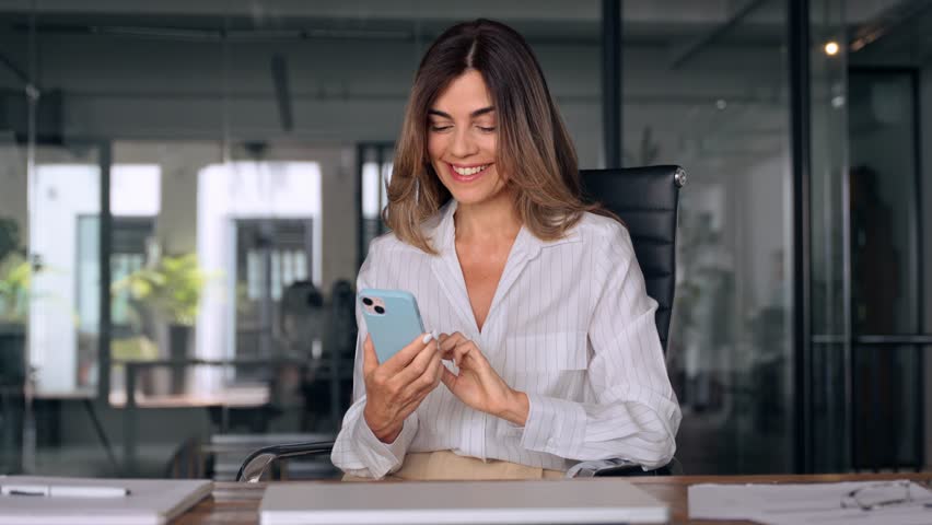 Smiling Latin Hispanic mature adult professional business woman using mobile phone cellphone. European businesswoman CEO holding smartphone using fintech application standing at workplace in office. | Shutterstock HD Video #1107342081