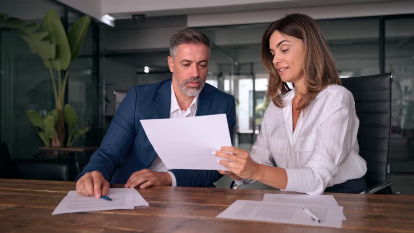 Mid age mature Latin businessman and European businesswomen discussing project with documents at table in office. Team of colleagues, diverse partners professional business people working together. Royalty-Free Stock Footage #1107342095