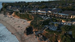 
The drone video captures a sunset flight starting from the beach, soaring over villa rooftops perched on cliffs, and gliding towards a golf course with the ocean beyond. 4k Aerial footage.