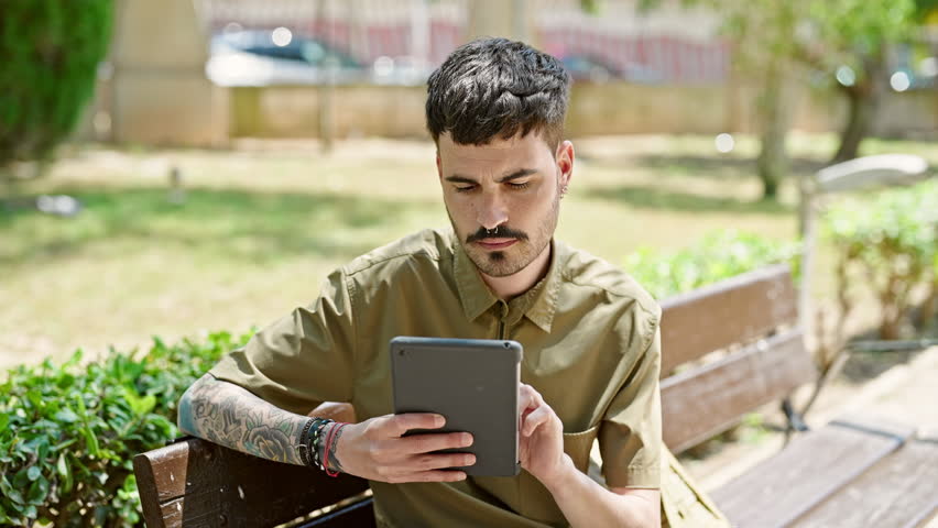 Young hispanic man tourist using touchpad sitting on bench celebrating at park