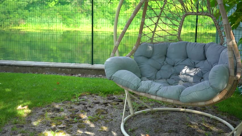 Ralaxing swing chiar on green summer lawn. Backyard cocoon basket made of weaving and grey mattress. Relax, cozy weeked near river, lake. Hanging bench seat. Relaxing in villiage house, hotel concept