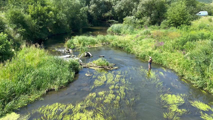 Aerial view of a beautiful river with clear flowing water, on which a naked fisherman in a white hat and underpants stands in the water, casting a spinning rod to catch predatory fish on a hot summer 