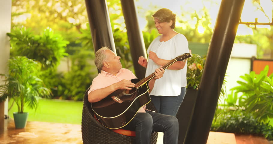 Happy mature middle aged man guitarist sitting on chair play acoustic guitar enjoy music outdoor home. Beautiful smiling Indian female singing practice tuning on musical instrument spend time together Royalty-Free Stock Footage #1107366163