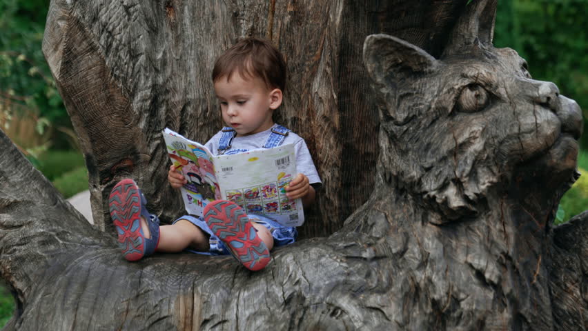 Serious baby boy sitting on top of a wooden cat sculpture. Adorable kid turns the pages of a children book.