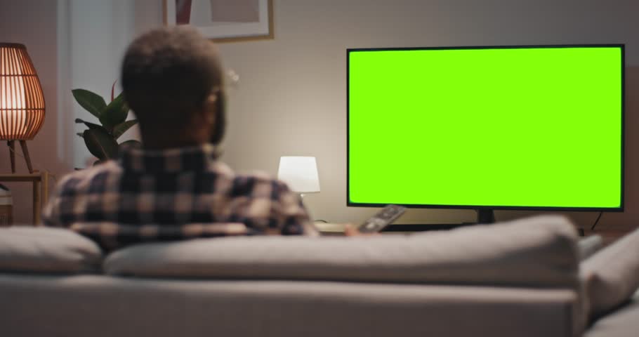 Man sitting at sofa and enjoying favourite TV show. Back view of male holding remote control and changing channel. Man relaxing sitting at couch at home. Male looking at green chroma key screen of TV. Royalty-Free Stock Footage #1107373279
