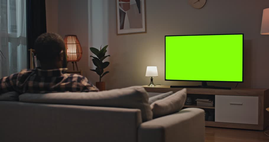Man sitting at sofa in modern appartment. Back view of male sitting in front of TV. African American man relaxing sitting at couch at well designed room. Male looking at green chroma key screen of TV. Royalty-Free Stock Footage #1107373281