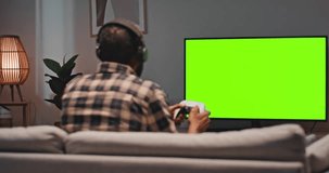 Man sitting at sofa and playing video games. Male holding joystick and wearing gaming headphones. Man relaxing sitting at couch at home. Male looking at green chroma key screen of TV.