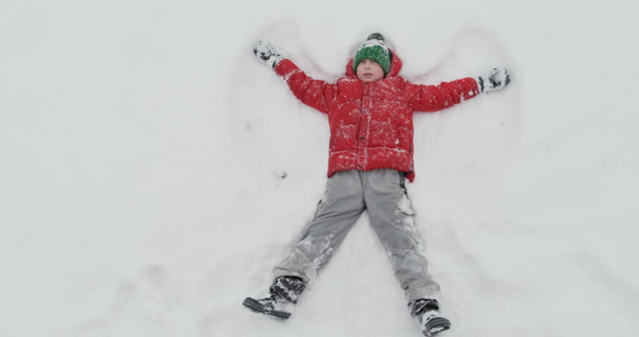 Boy in red jacket lies in snow and makes snow angel. Top view. Entertainment for children in winter.