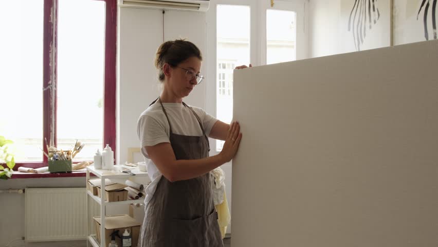 The artist checks how well the new canvas is stretched on the frame and carries it to put against the wall and start new work. The artist carries a large, blank canvas. Artist's workshop. Royalty-Free Stock Footage #1107402825