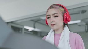 Young Asian woman in headphones video call with friends showing muscles while exercising on treadmill
