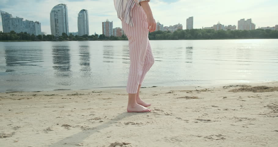 Young woman legs walk, sand on river beach. Wide river, skyscrapers of city are reflected on surface of water. Gimbal follow, side view