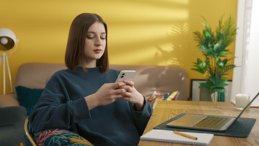 Smiling girl texting message on smartphone. Female student taking rest between online lessons at home. Attractive woman using mobile phone. Young lady posing at the camera in room, 4k footage