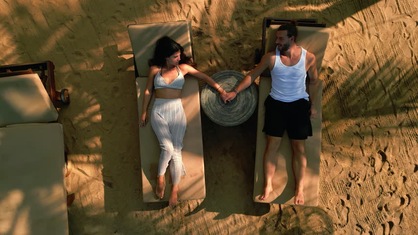 Multiracial couple on sunbeds by the sea hold hands top view from a drone. Young biethnic man and woman chill on picturescue tropical sandy ocean beach under palm trees aerial ascending shot.