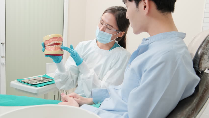 Asian female dentist explaining orthodontic care and whitening to young male patient with teeth model in dental clinic, well-being hygiene, and professional healthcare work in doctor office hospital. Royalty-Free Stock Footage #1107420225
