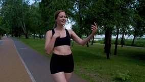 Positive attractive fitness woman in activewear live streaming video using cellphone, communicating and sharing with audience online about active lifestyle while walking along park after workout.