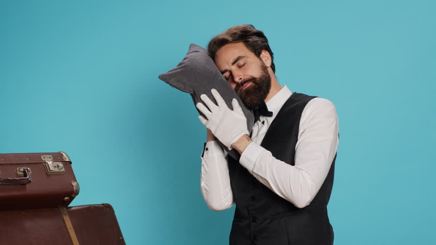 Sleepy bellman posing with pillow in studio, advertising burnout and being overworked in classy hospital industry. Hotel porter taking quick nap after working overtime, tired doorman. Royalty-Free Stock Footage #1107432043