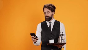 Catering employee texting messages on phone while carrying restaurant tray with drinks and cutlery, posing in studio. Sophisticated waiter in suit and tie looking at smartphone display. - Powered by Shutterstock - Get 15% off with code: PIKWIZARD15