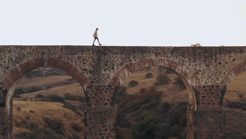 ariel shot of a fashionable and dearing man walking across a small bridge in italy on a hot summers day in the afternoon.