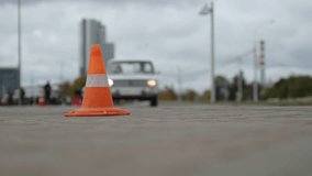 Orange traffic cone is set on road in city, ensuring safety after traffic accident. Old car with its headlights on drives up to cone and turns on turn signal to avoid obstacle on cloudy day. - Powered by Shutterstock - Get 15% off with code: PIKWIZARD15