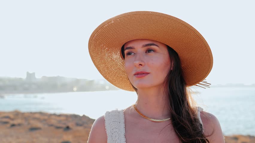 Pretty young Caucasian woman smiling looking at the camera. Cute girl in a hat standing by the sea on a beach, enjoying a summer vacation. Stunning lovely young lady spending time on journey.