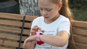 Press the display on the wrist children's smart watch. Internet on the clock. A little girl with a clock in her hands sits in the park and looks closely at the display. Gadget addiction