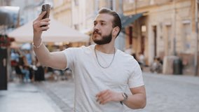 Bearded young man blogger taking selfie on smartphone, communicating video call online with subscribers, recording stories for social media vlog outdoors. Guy walking in urban sunshine city street