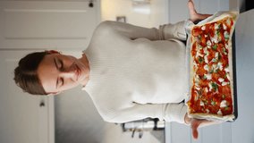 Vertical video of young smiling woman in kitchen at home holding fresh pizza ready for the oven - shot in slow motion