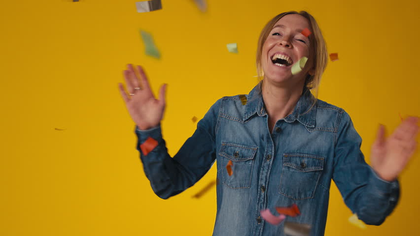 Studio shot of excited woman celebrating big win showered in tinsel confetti on yellow background - shot in slow motion Royalty-Free Stock Footage #1107475817